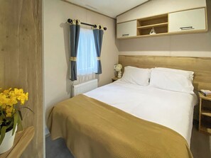 Habitación de hotel con cama, mesita de noche, estante con un objeto pequeño, ventana con cortinas y una planta en maceta.