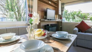 Un coin repas avec une table en bois, des bols, un saladier en verre rempli de fruits, un vase avec des fleurs et, au fond, un téléviseur.