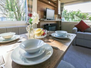 Un comedor con una mesa de madera, tazones, un tazón de vidrio con fruta, un florero con flores y un televisor al fondo.