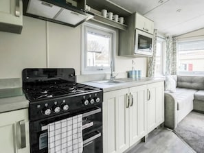A compact kitchen with a stove, oven, and sink. There is a window above the sink and a television mounted above the oven. - Goscar 1 The Saltern Holiday Park - UKC8209 (Tenby)