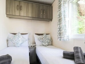 A bedroom with two beds, a wooden wardrobe, a window with a floral curtain, and a 'love' sign on the wall. - Goscar 1 The Saltern Holiday Park - UKC8209 (Tenby)