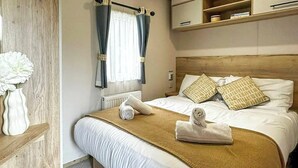A hotel room with a bed, pillows, a bedside table, a vase with flowers, and a window with curtains. - Coastal 1 The Saltern Holiday Park - UKC8200 (Tenby)