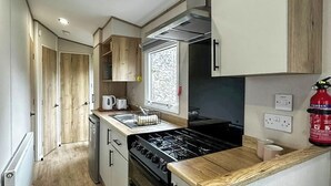 A compact kitchen with a stove, oven, and sink, wooden cabinets, and a fire extinguisher. - Coastal 1 The Saltern Holiday Park - UKC8200 (Tenby)