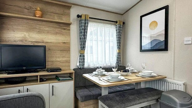 A compact room with a television, a dining table set for two, a bench, and a trash bin. - Coastal 1 The Saltern Holiday Park - UKC8200 (Tenby)