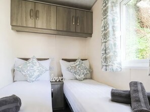 A bedroom with two beds, a wardrobe, a window with a curtain, and a small bedside table. - Goscar 2 The Saltern Holiday Park - UKC8210 (Tenby)