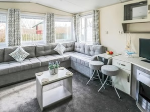 A modern living room with a grey sectional sofa, a small white table, and a kitchen area with a TV and a sailboat model. - Tenby 4 The Saltern Holiday Park - UKC8207 (Tenby)