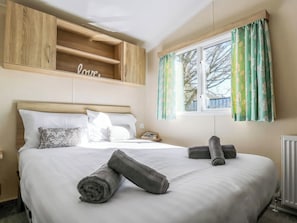 A neatly made bed with pillows, a wooden shelf above, and a window with curtains. - Tenby 4 The Saltern Holiday Park - UKC8207 (Tenby)