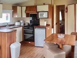 A compact kitchen with a sink, stove, microwave, and a small refrigerator. There is a dining table with chairs and a trash bin. - Coastal 4 The Saltern Holiday Park - UKC8203 (Tenby)