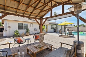 Un patio techado al aire libre con mobiliario de madera, una pila de bloques de madera y una zona de piscina con sillas y una sombrilla.