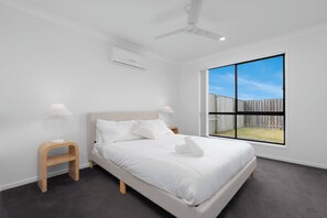 A modern bedroom with a bed, two bedside tables, a ceiling fan, and a large window with a view of a fenced yard. - Scandi Calm - Sleek 1BD Duplex near Everything (Pimpama)