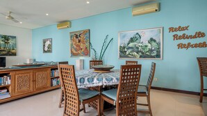 Ruokailutilassa on neljälle katettu pöytä, ja sen ympärillä on puisia tuoleja, joiden selkänojissa on koristeellinen ristikko. Seinällä on kaksi maalausta, joista toisessa on kuvattuna trooppinen maisema ja toisessa kukkakimppu. Pöydällä on koristeellinen kulho, ja vasemmalla puolella on kirjahylly, joka on täynnä kirjoja.