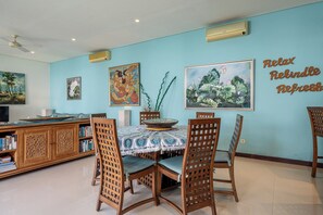 A dining area with a table set for four, surrounded by wooden chairs with lattice backs. There are two paintings on the wall, one depicting a tropical scene and the other a floral arrangement. A decorative bowl is on the table, and there is a bookshelf filled with books to the left. - Villa Samsara – 3BR Pool Villa Near Berawa Beach (Kabupaten Badung)