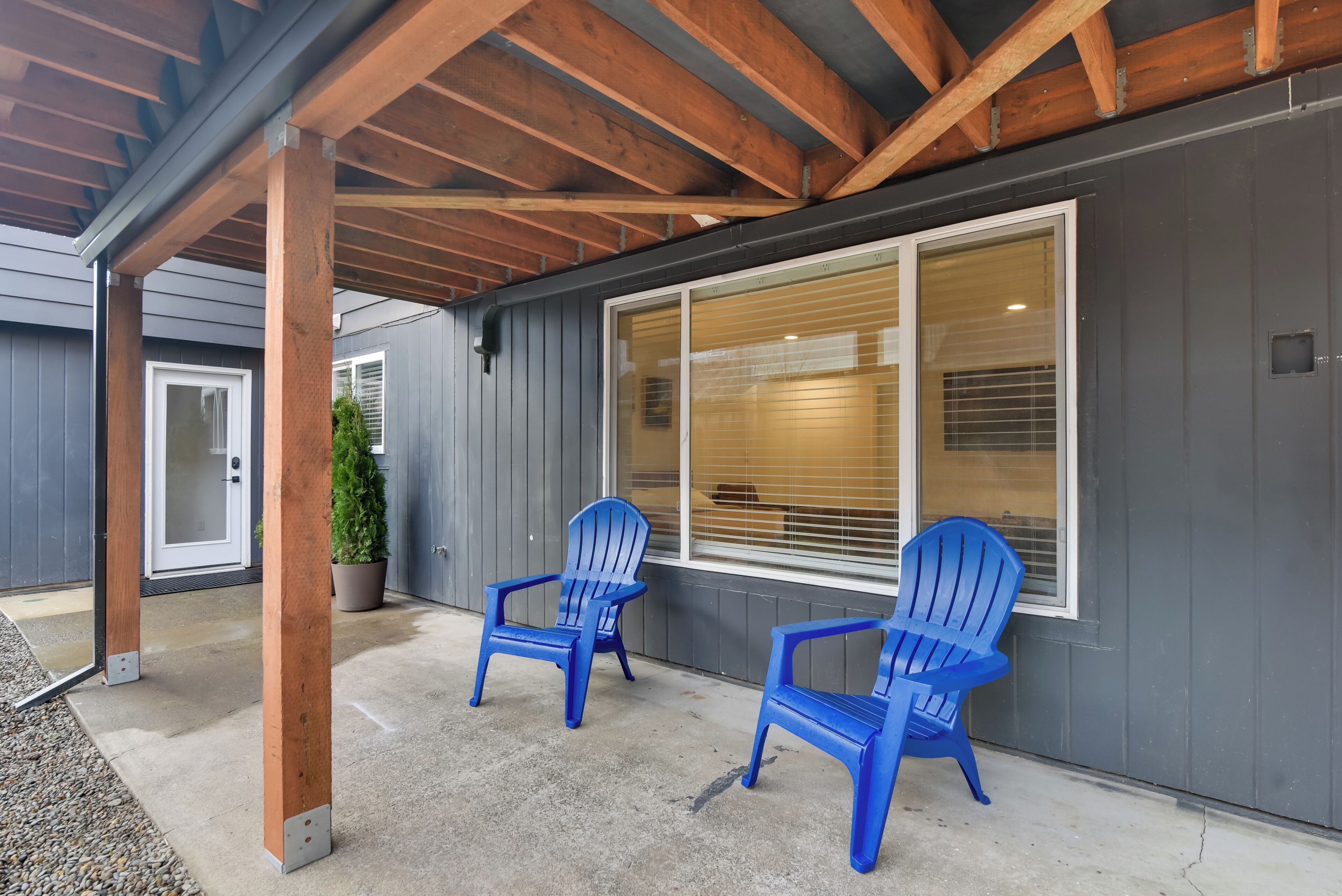 A covered porch with two blue Adirondack chairs, a potted plant, and a large window with blinds.