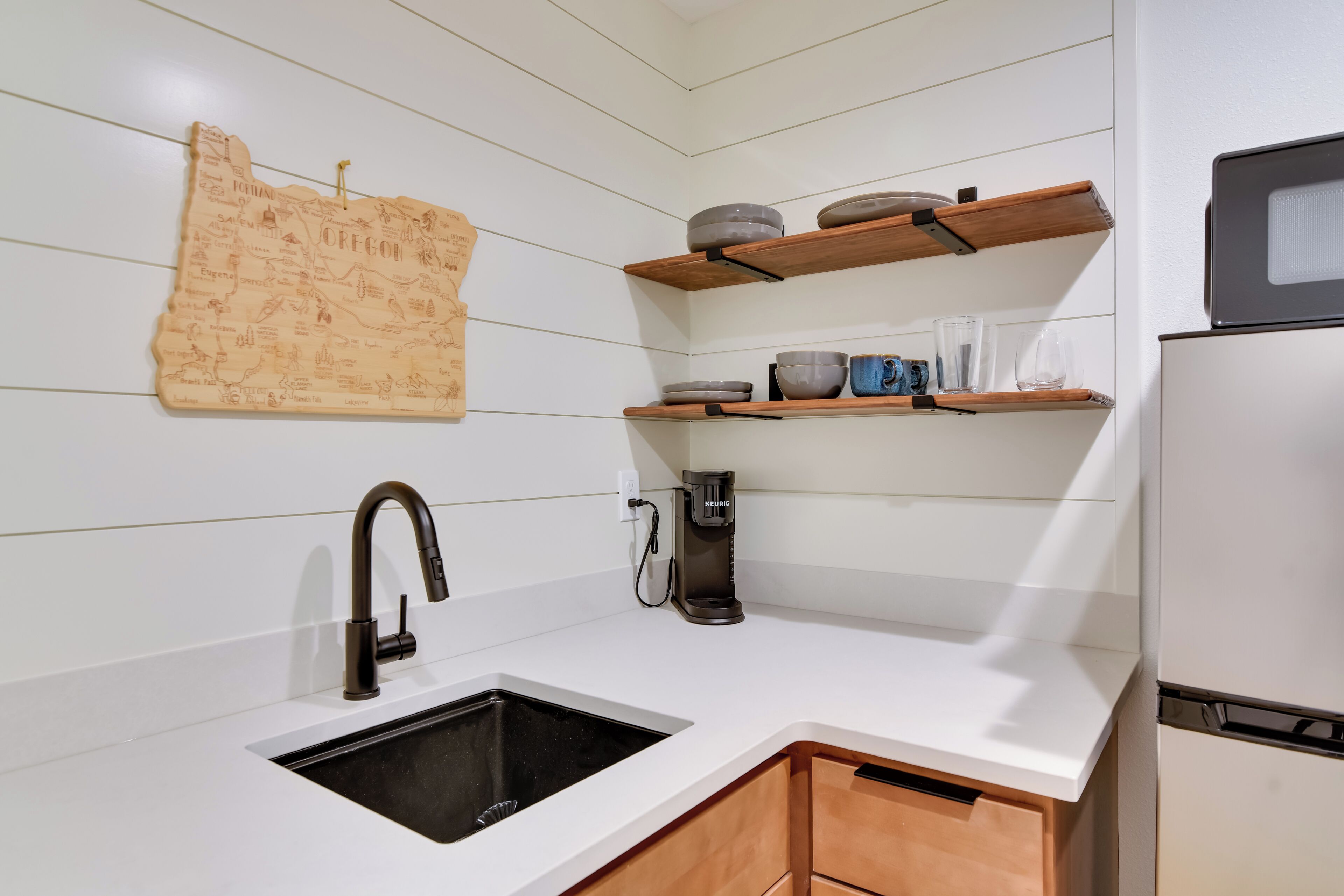 Une cuisine avec un plan de travail blanc, un évier noir, des étagères en bois où sont rangés de la vaisselle, une machine à café et une carte accrochée au mur.