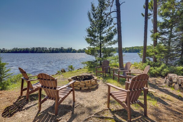 A lakeside setting with wooden Adirondack chairs arranged around a fire pit, surrounded by trees and a clear view of the lake. - Private Peninsula! Laurel Lake Retreat w/ 3 Piers (Three Lakes)