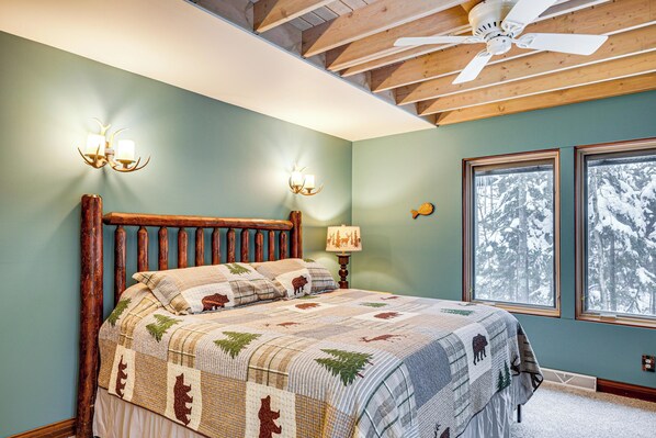 A bedroom with a wooden bed, a quilt with animal prints, two wall sconces, a ceiling fan, and a window with a snowy view. - Private Peninsula! Laurel Lake Retreat w/ 3 Piers (Three Lakes)