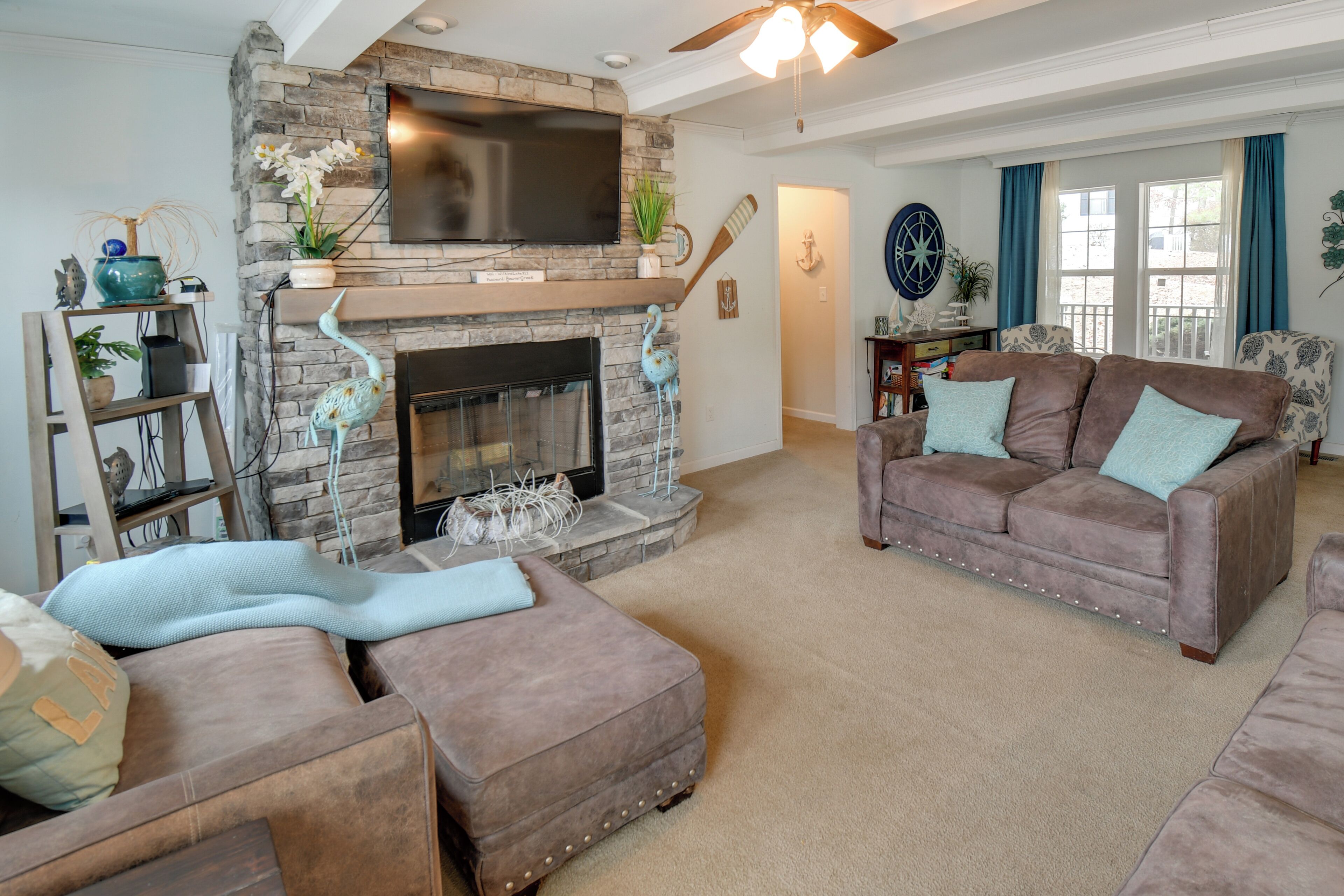 A living room with a stone fireplace, a flat-screen TV mounted above it, and a comfortable seating area with a brown sofa and ottoman.
