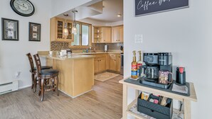 Una cocina moderna con armarios de madera, una isla central con taburetes de bar, una barra para café con máquina Keurig y un reloj en la pared.