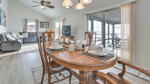 Un comedor con una mesa de madera dispuesta para cuatro personas, rodeada de sillas de madera. Se observan un ventilador de techo y luces colgantes sobre la mesa.