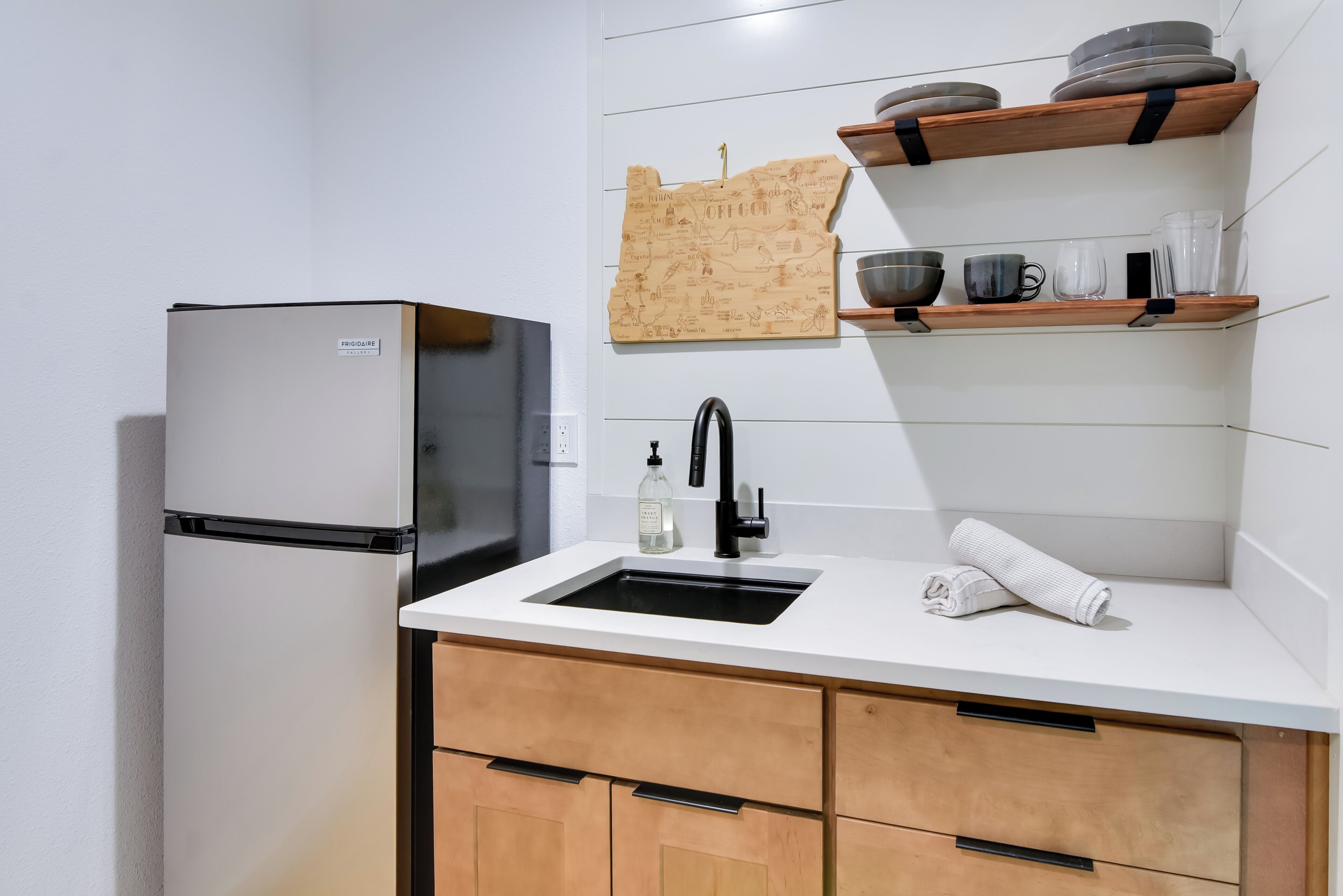Une cuisine équipée d’un réfrigérateur, d’un évier, de meubles de rangement en bois et d’étagères où sont rangées la vaisselle et les verres.