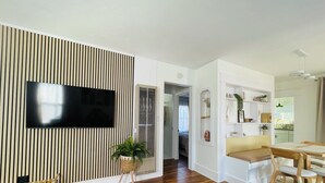 Una sala moderna con un televisor de pantalla plana montado en una pared a rayas, un banco de madera, un reposapiés verde y una zona de comedor con una mesa y sillas de madera.