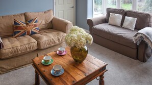 A living room with two sofas, a wooden coffee table with cups and a vase, and a view of a garden through large windows. - 3 bedroom accommodation in Sheringham (Sheringham)