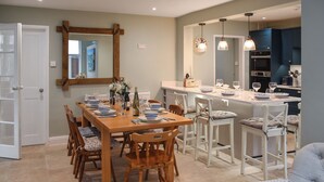 A dining area with a wooden table set for six, surrounded by white chairs with cushioned seats. A kitchen area with modern appliances and pendant lighting is visible in the background. - 3 bedroom accommodation in Sheringham (Sheringham)