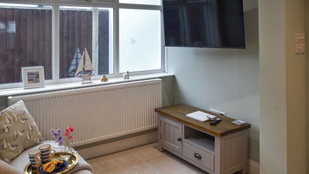 A cozy living room with a sofa, a small wooden table, a radiator, and a television mounted on the wall. - 3 bedroom accommodation in Sheringham (Sheringham)