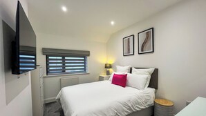 Ein modernes Schlafzimmer mit einem Bett, einem an der Wand montierten Fernseher, einem Fenster mit Jalousien, einer Nachttischlampe und zwei gerahmten Bildern an der Wand.