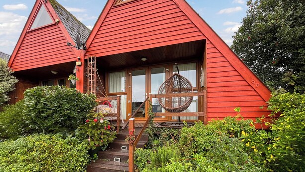 Et rødt, trekantet hus med en trætrappe, der fører op til en veranda, en hængestol og nummer '95' på siden.