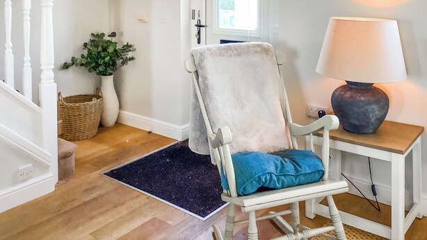 Ein Zimmer mit Holzboden, einem weißen Schaukelstuhl mit blauer Kissen, einem kleinen Tisch mit Lampe, einem Korb und einer Zimmerpflanze.