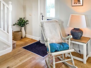 Una habitación con piso de madera, una mecedora blanca con cojín azul, una pequeña mesa con lámpara, una cesta y una planta en maceta.