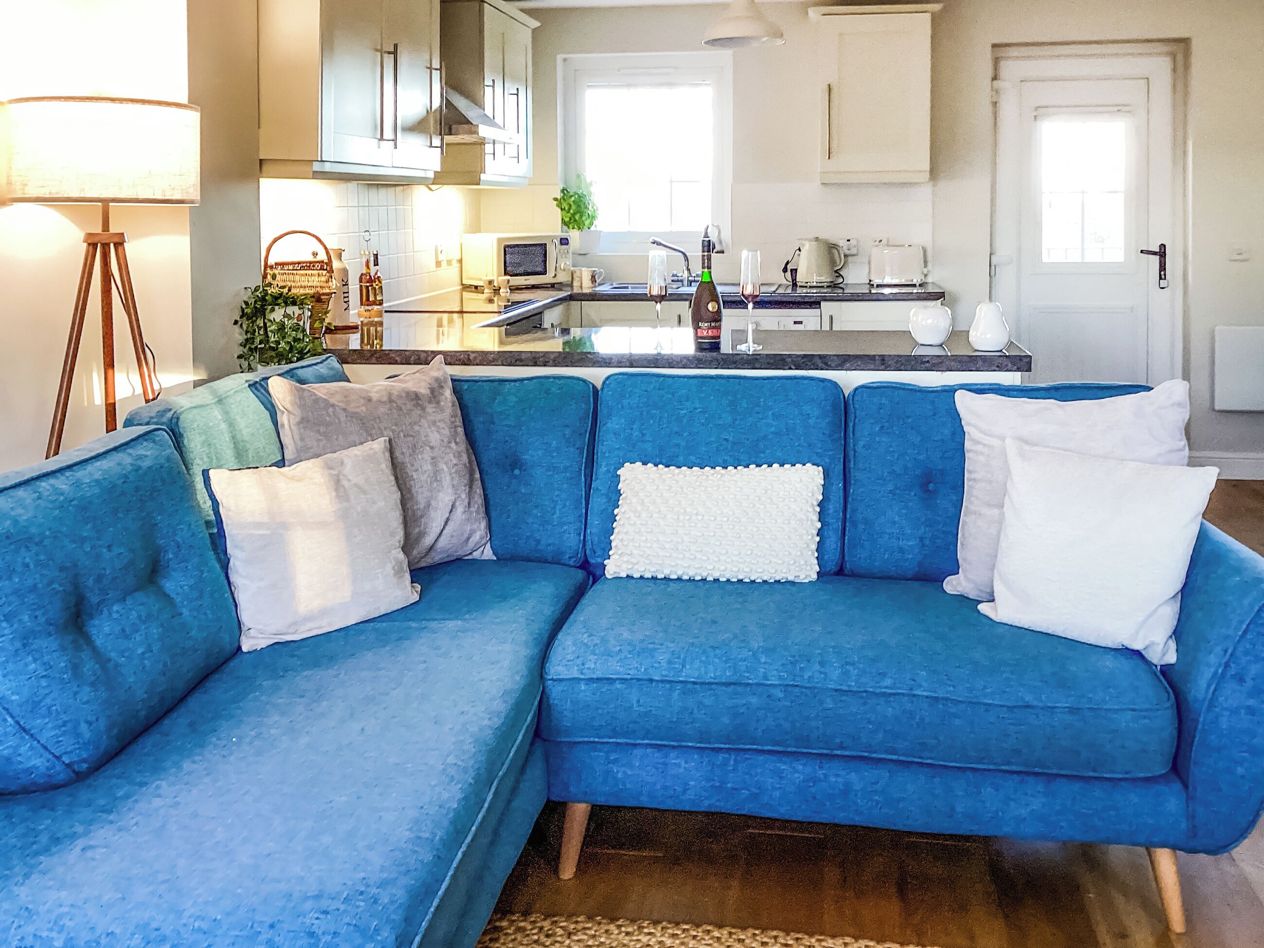 En moderne stue med en blå sofa, et køkkenafsnit med mikroovn, brødrister og vask, samt en dør til et andet rum.