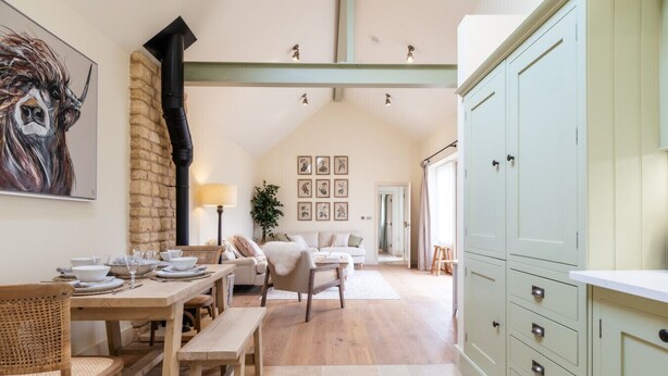 Un comedor con una mesa y sillas de madera, una pared de piedra y un cuadro grande. Se ve una cocina con armarios verde claro y una encimera blanca.