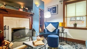 Una habitación con chimenea, un sillón azul, una mesa redonda de cristal con un libro, una mesita auxiliar con una lámpara y una ventana con vista a un edificio de ladrillo.