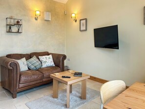 Una sala de estar con un sofá de cuero marrón, una mesa de centro de madera, un televisor montado en la pared y un estante pequeño con adornos.