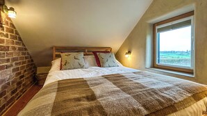 Ein Schlafzimmer mit schrägverdeckter Decke, einer Ziegelwand, einem großen Fenster und einem Bett mit gemusterten Betttextilien.