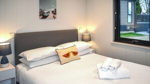 Cama bem arrumada com lençóis brancos, um travesseiro com desenho de pássaro, uma luminária na mesa de cabeceira e uma janela com vista para uma casa e gramado.