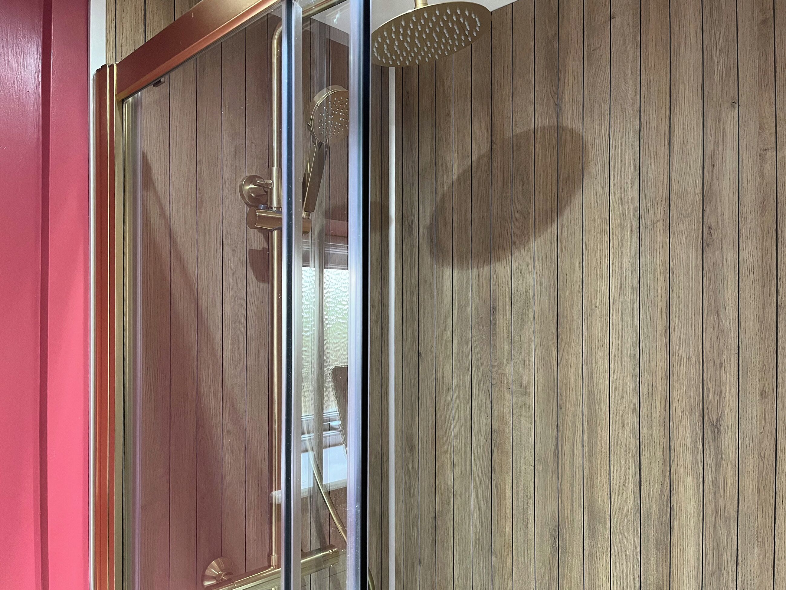 A shower with a glass door, a wooden wall, and a metal showerhead.