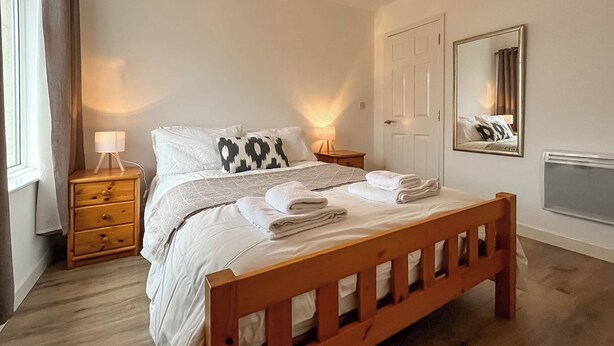 A bedroom with a wooden bed, white bedding, two bedside tables with lamps, a mirror, and a door.
