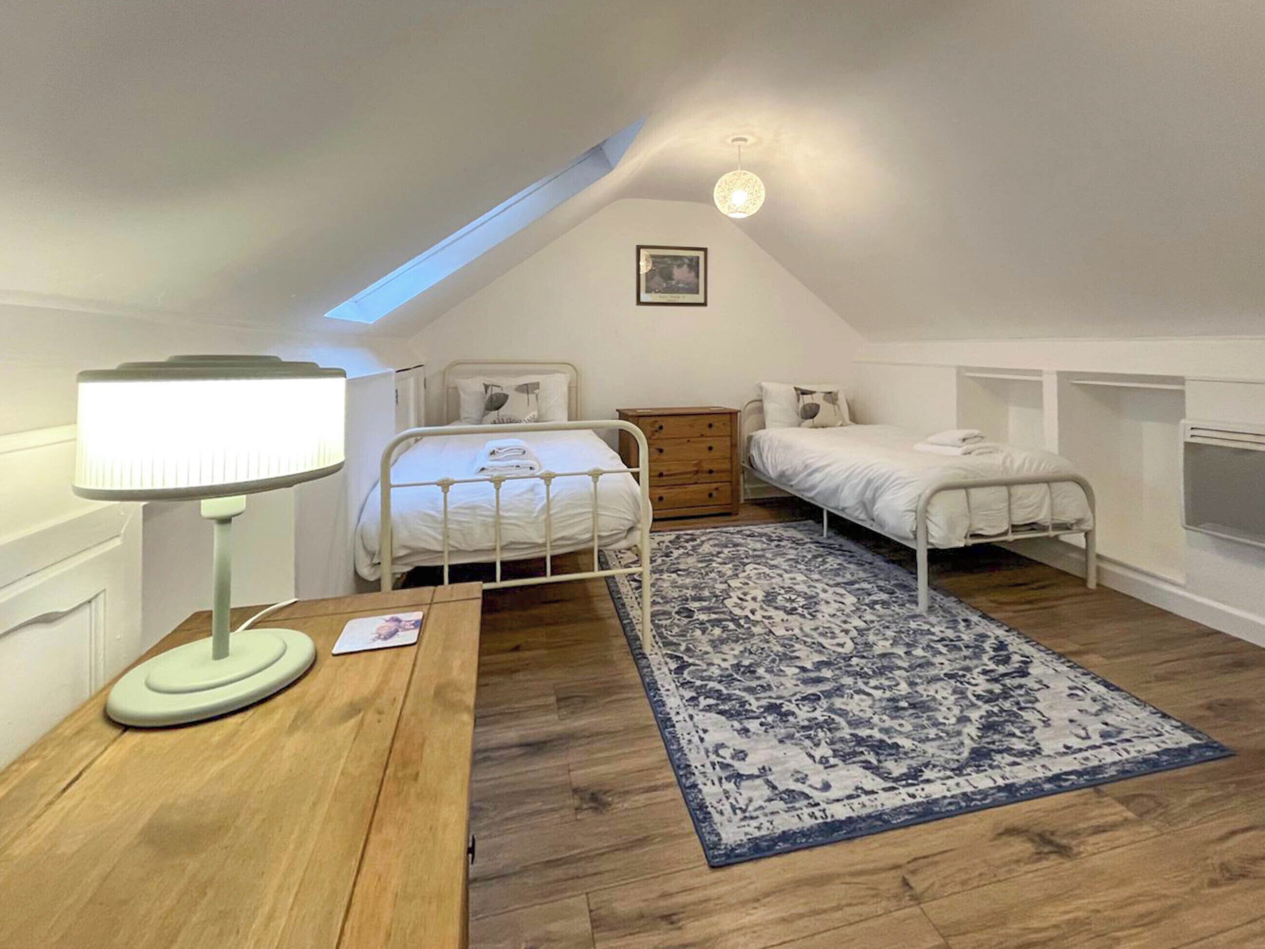 A small, white, attic-style bedroom with two beds, a wooden desk, a lamp, and a patterned rug.