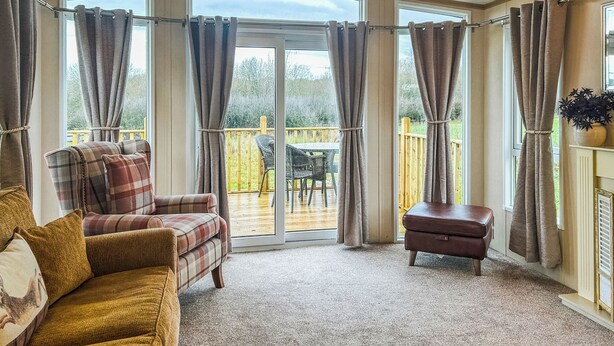 A cozy living room with a sofa, armchair, and ottoman, large windows with curtains, and a view of a deck. - 2 bedroom accommodation in Hartshorne (Hartshorne)