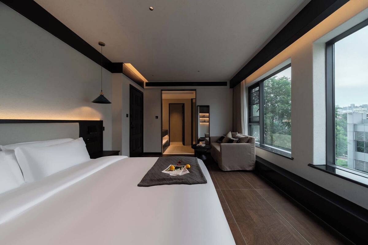 A modern hotel room with a large bed, a seating area with a chair, and a view of the city through large windows.