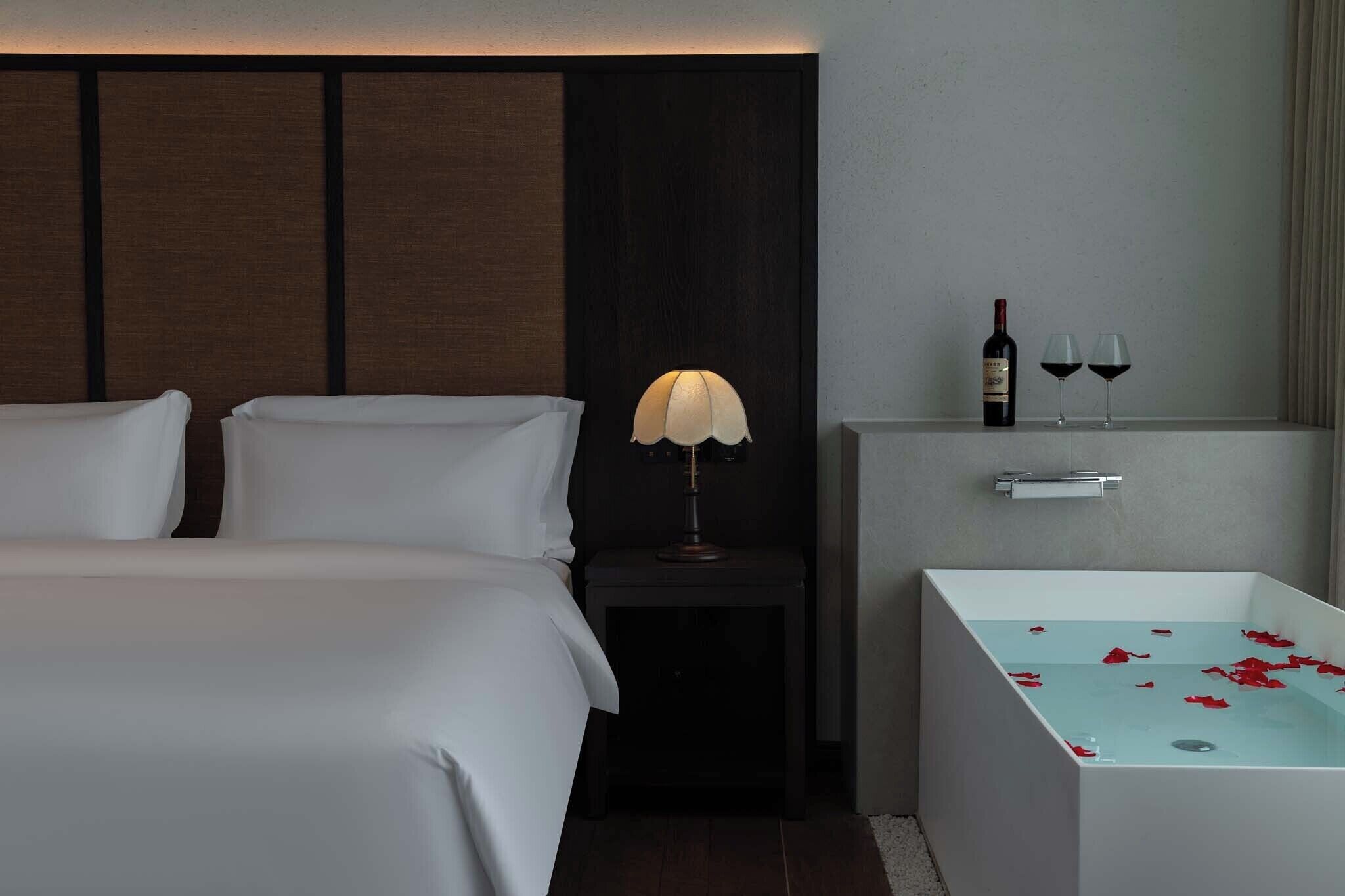A hotel room with a bed, a nightstand with a lamp, a minibar, and a bathtub with rose petals.