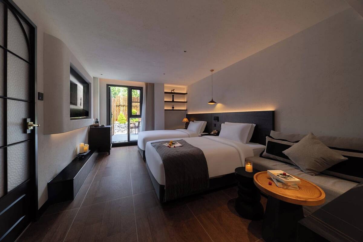 A modern hotel room with two beds, a sofa, a small table, and a view of greenery outside.