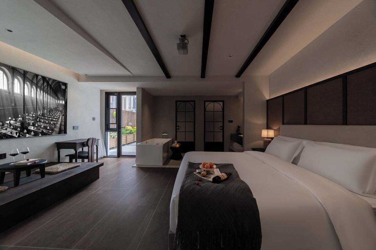 A modern hotel room with a large bed, a seating area with a table and chairs, a bathtub, and a large window with a view of the outside.