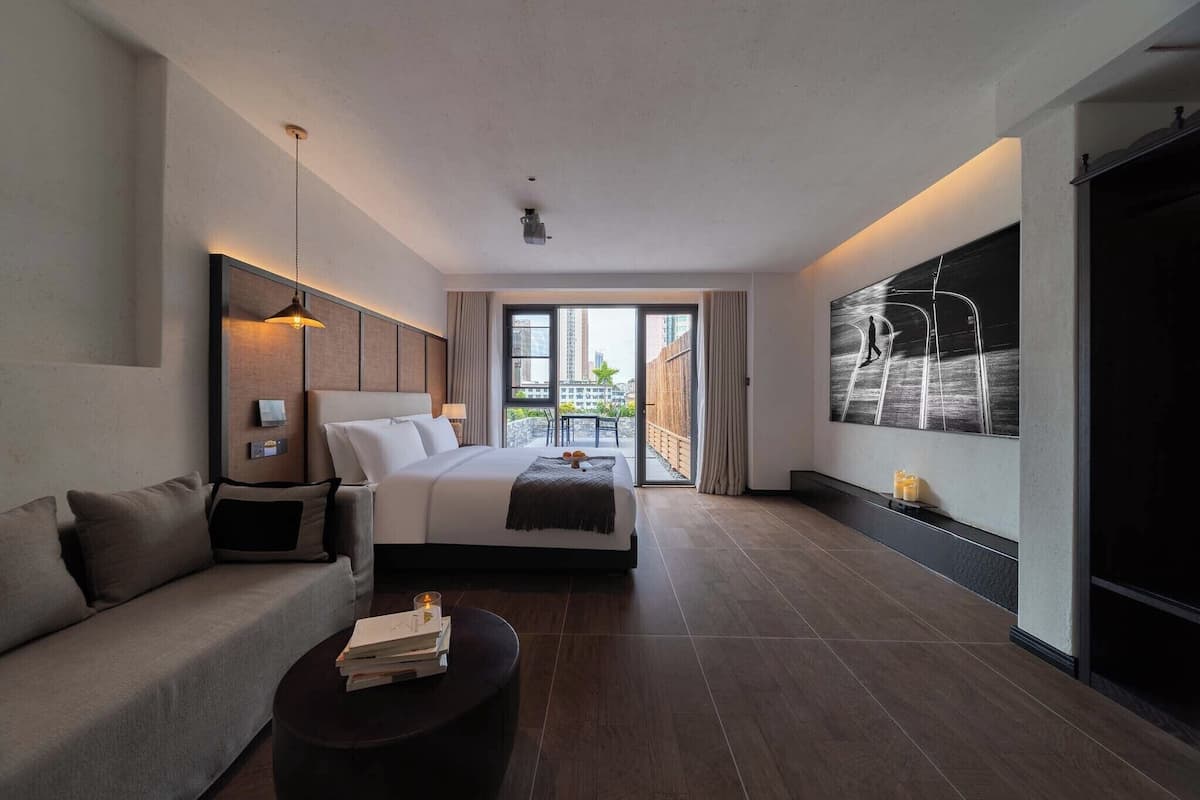 A modern hotel room with a large bed, a sofa, a coffee table, and a view of the city through large windows.