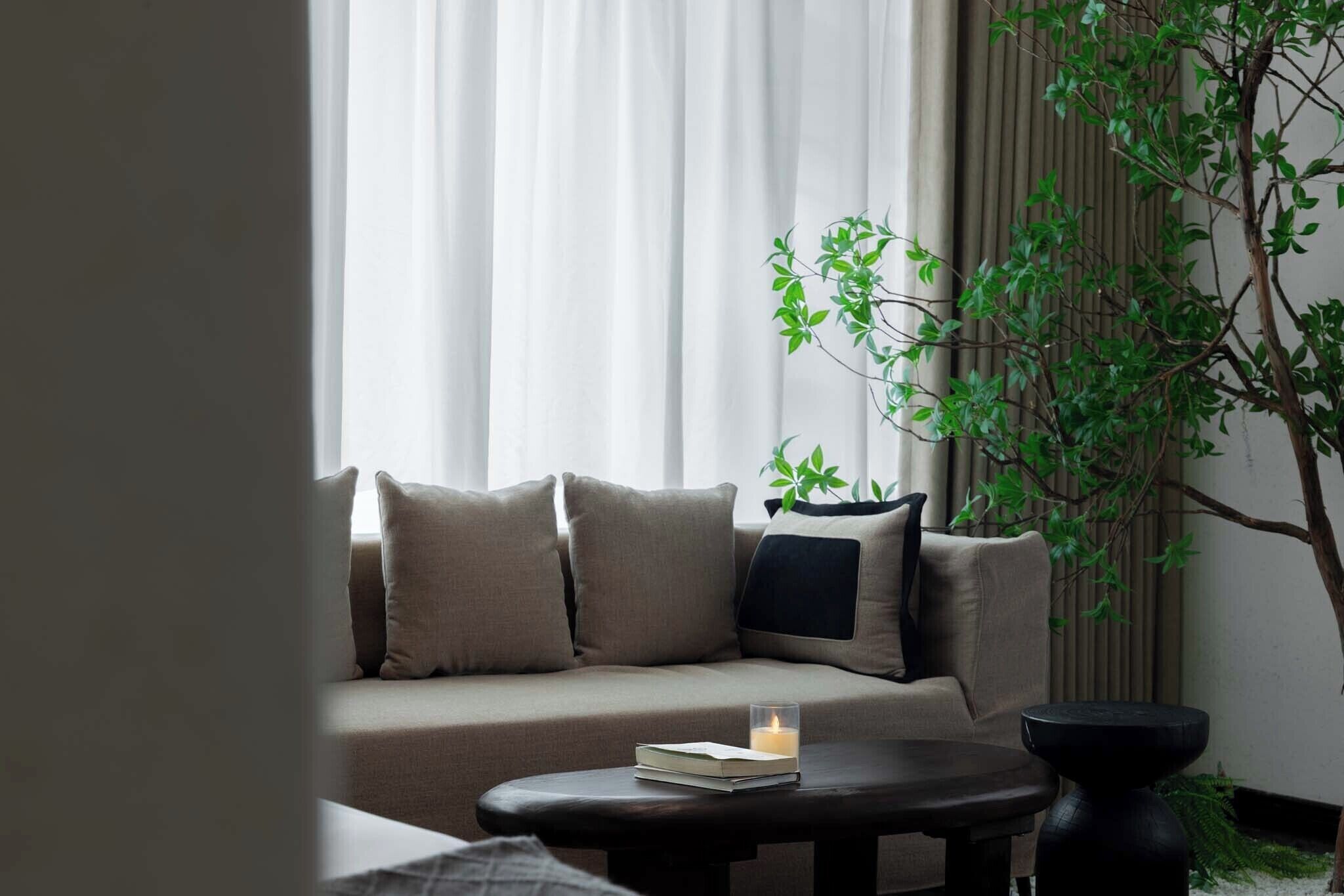 Una sala de estar moderna con un sofá beige, una mesita redonda con un libro y una vela, y una planta en maceta.