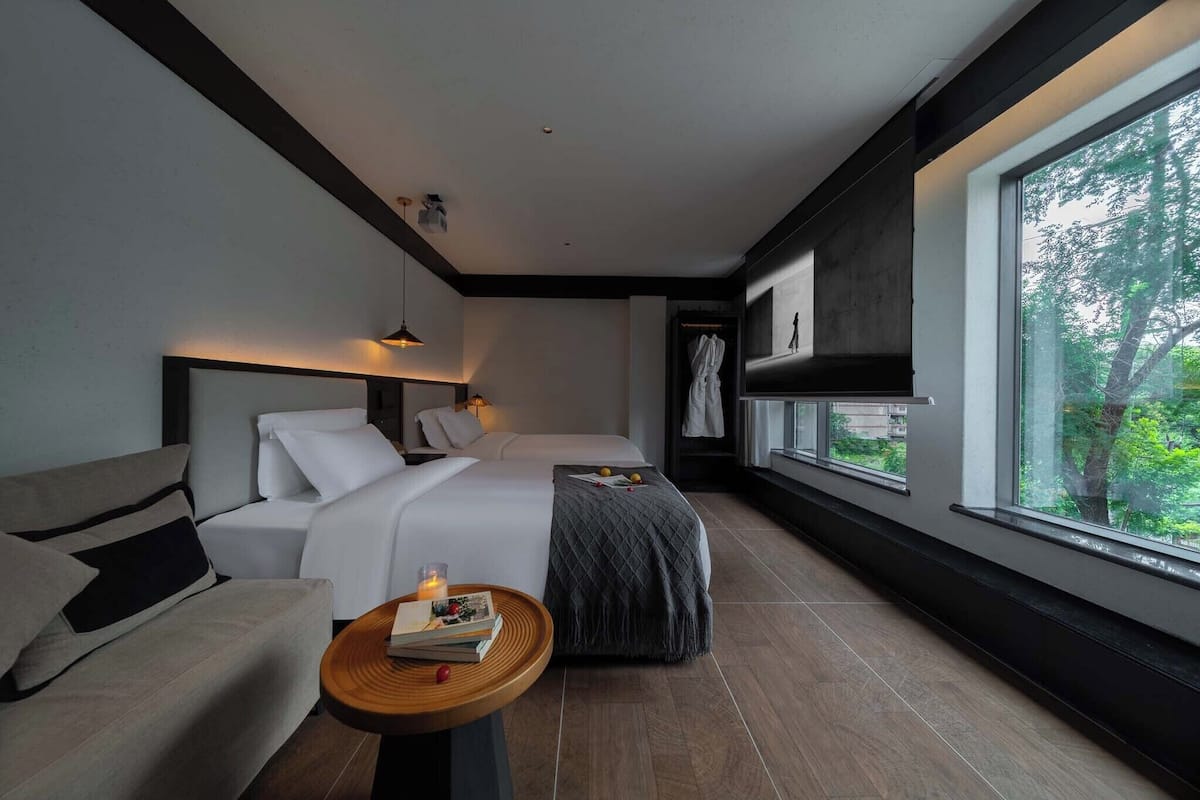 A modern hotel room with a bed, a sofa, a small table with a candle, and a large window offering a view of trees.