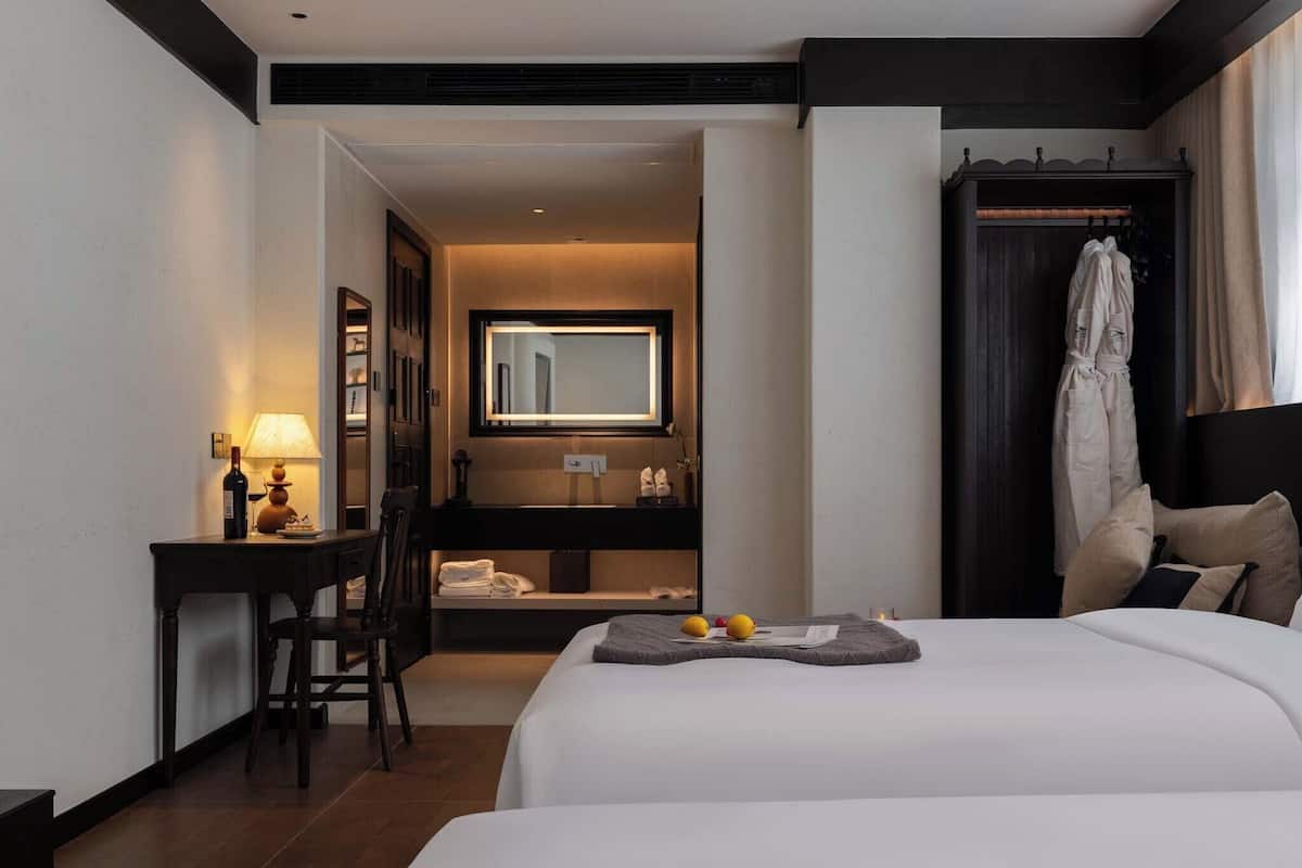 A hotel room with a bed, a desk with a lamp, a mirror, a robe hanging, and a small table with fruits.